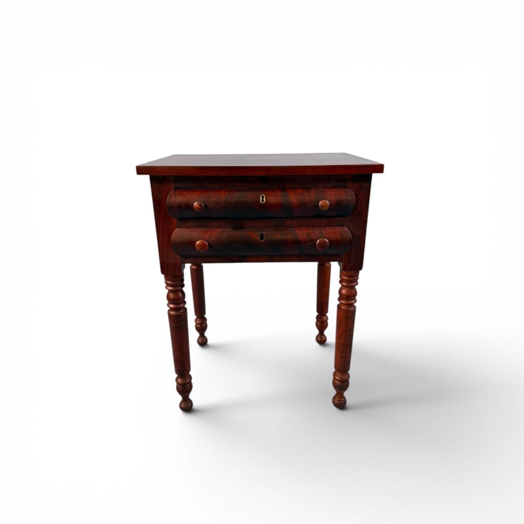 American Two-Drawer Work Table in Figured Mahogany: American work table in figured mahogany, the rectangular top above two aligned drawers with turned wooden pulls and escutcheons, raised on turned legs. Drawer fronts display strong grain patterning;