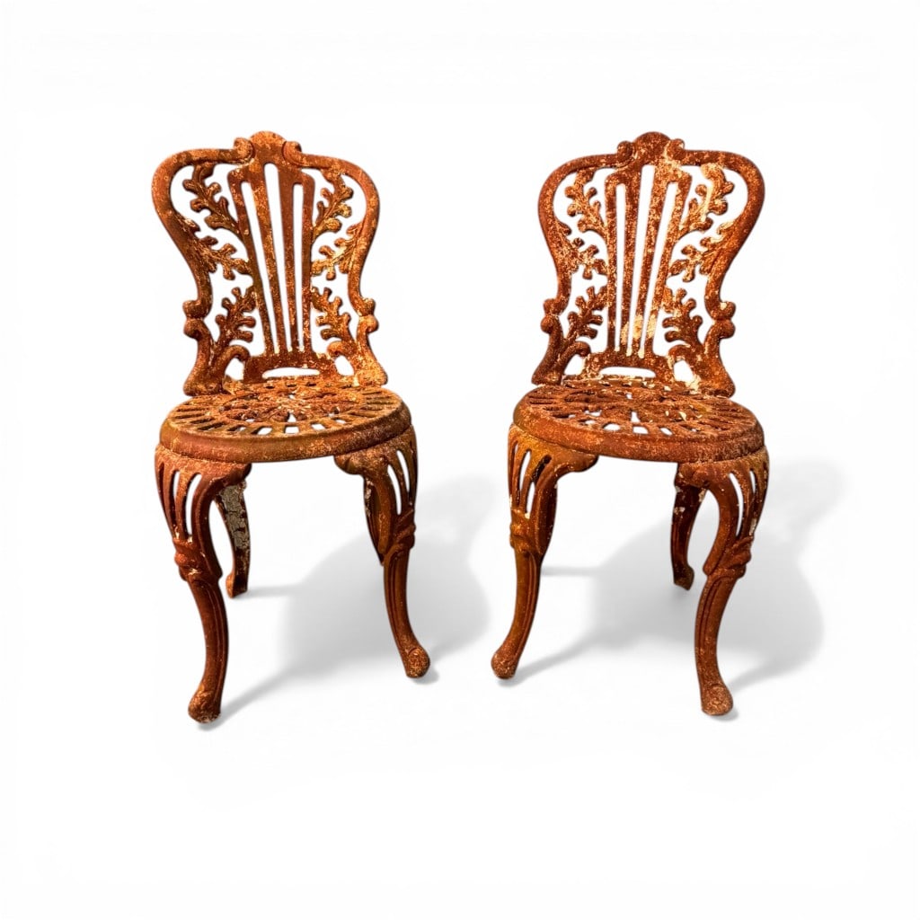 Pair of Cast Iron Garden Chairs with Foliate Scrollwork: A decorative pair of cast iron garden chairs featuring pierced backs with stylized foliate and scrollwork ornament, centered by a vertical splat motif. The circular seats display a radiating openwork