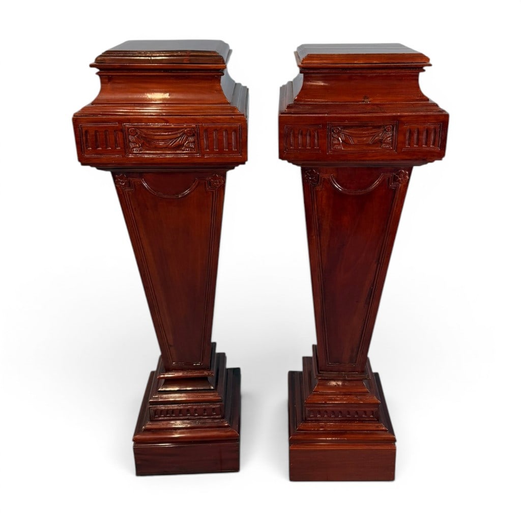 Pair of Neoclassical Mahogany Pedestals/Stands: Pair of neoclassical-style mahogany pedestal stands, each with square stepped top above a molded plinth-form capital, the frieze carved with classical drapery swags, flanking fluted panels, and