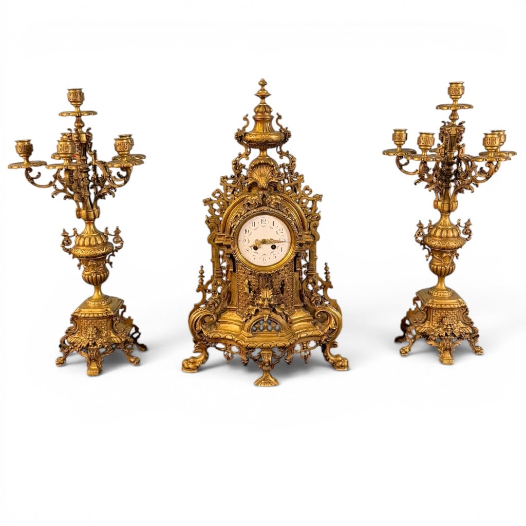 Three-Piece Gilt Brass Garniture Clock Set: Three-piece gilt brass garniture clock set, late 19th/early 20th century, in the French Rococo Revival taste. The clock with highly ornate cast case having urn finial, shell crest, pierced