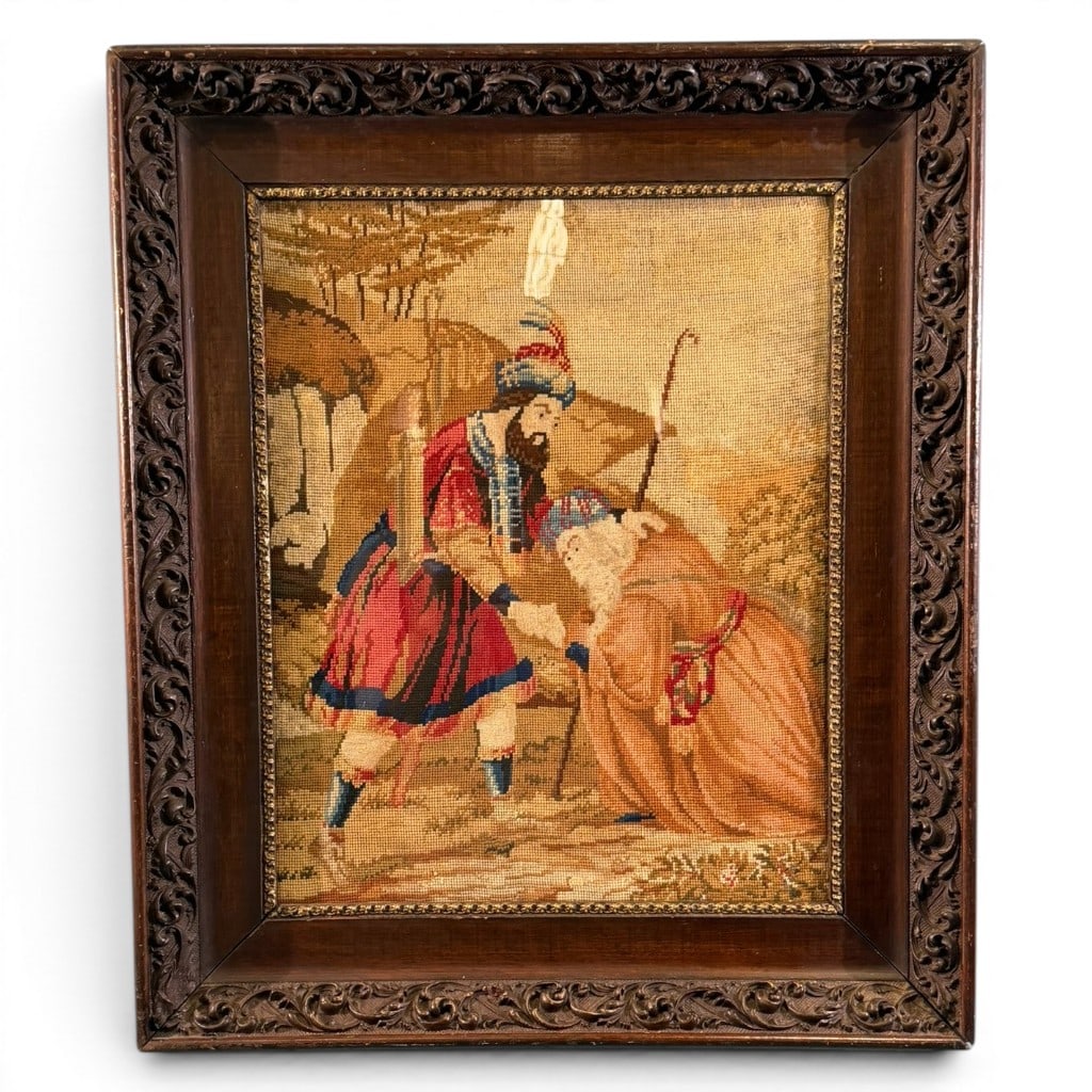 European Needlework Figural Scene: Framed European needlework panel depicting an Orientalist/Biblical-style figural scene with a robed male figure, kneeling animal, and landscape background, mounted under glass in a carved dark wood