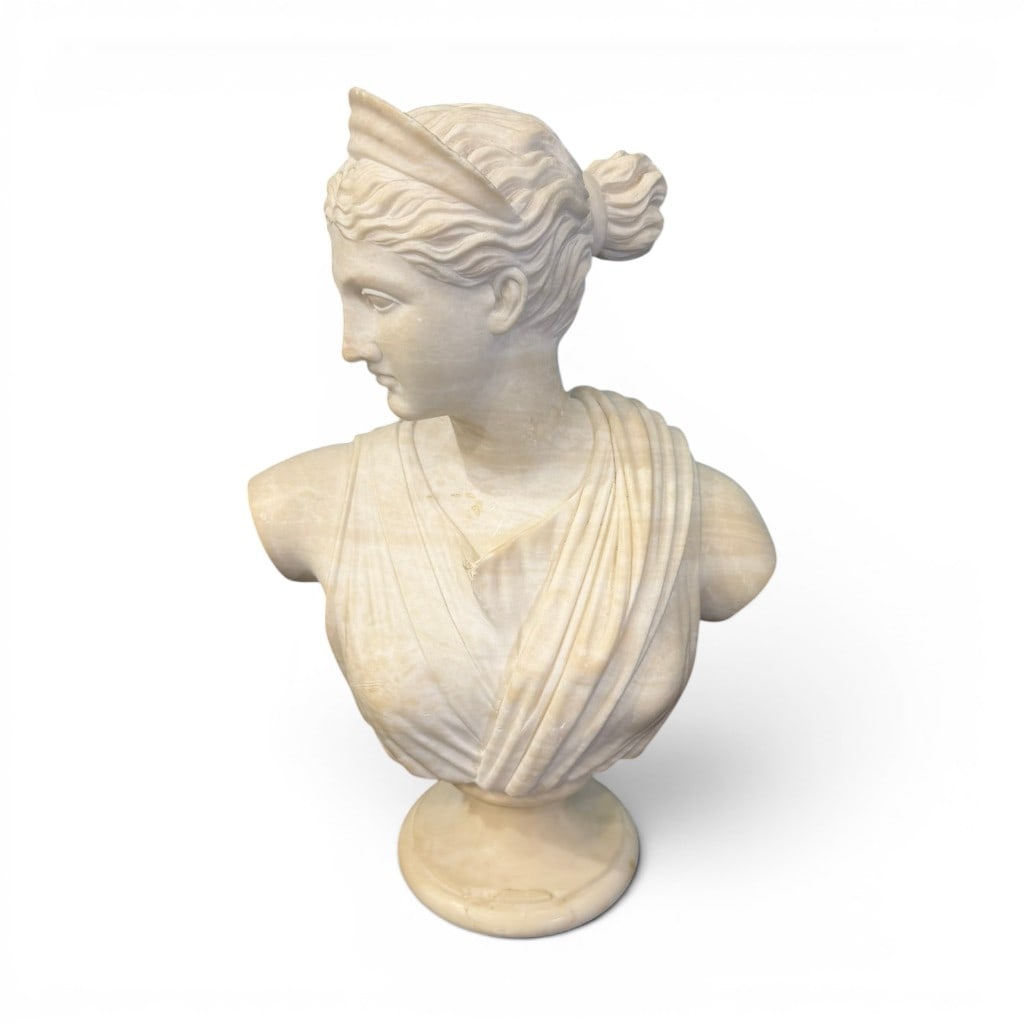 Carved Marble Bust of a Classical Female Figure, Diana or Athena: Fine carved marble bust of a classical female figure, likely depicting Diana or Athena, modeled with serene downward gaze, classical drapery, upswept hair, and wing-form or helmet-like head ornament,