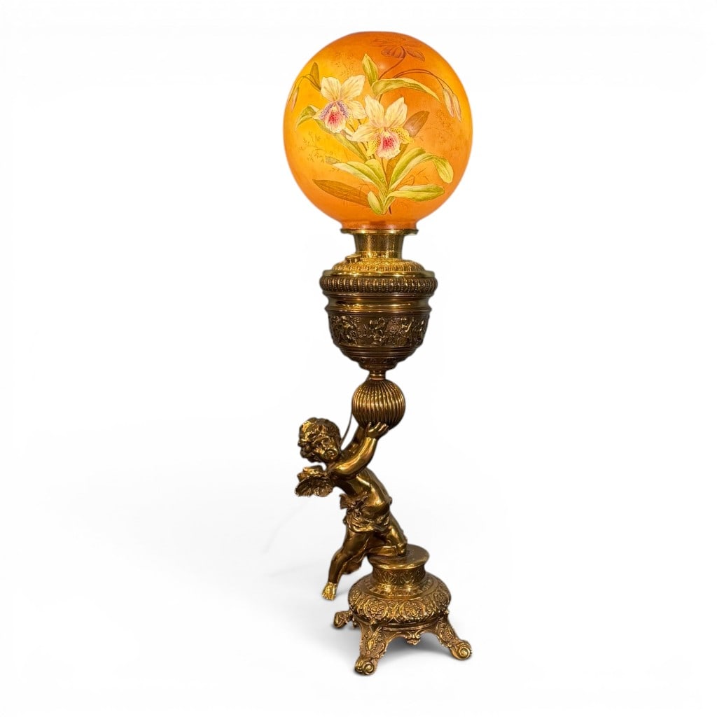 Victorian Figural Brass Parlor Lamp with Painted Glass Globe: A fine Victorian figural parlor lamp, late 19th century, having an ornate cast brass and gilt-metal base modeled as a winged putto or cherub supporting a ribbed sphere beneath an elaborate burner