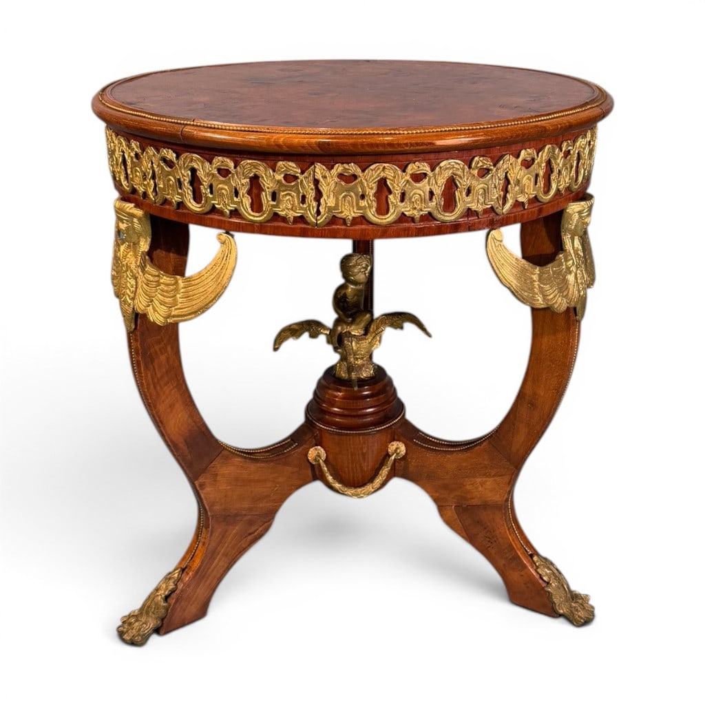 Egyptian Revival Gilt-Mounted Burlwood Center Table, late 19th century: A striking Egyptian Revival circular center table, late 19th century, the round burlwood top with beaded brass gallery edge above a deep apron mounted with gilt-metal anthemion, lotus, and interlaced