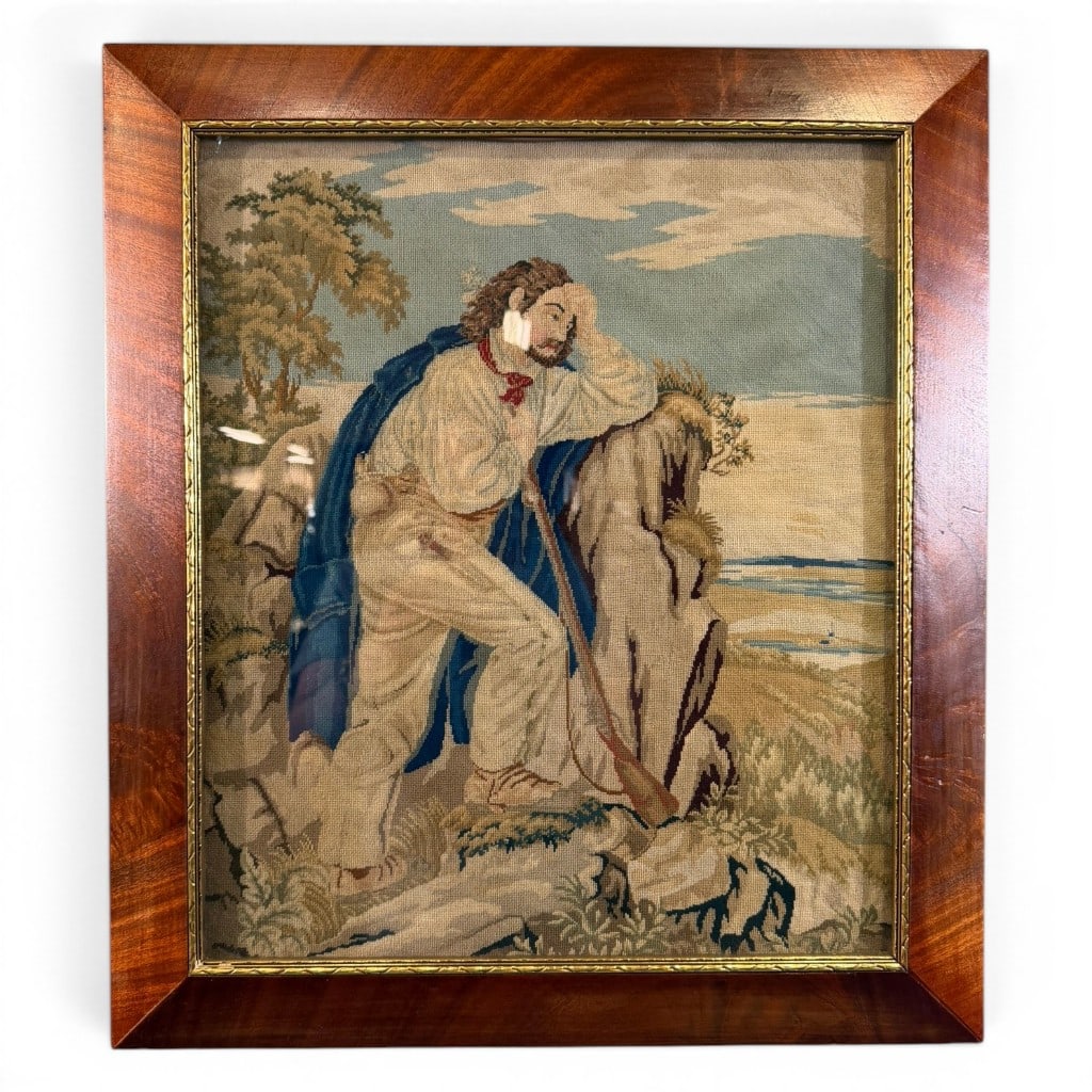 Needlepoint Soldier or Traveler Scene: Framed needlepoint panel depicting a soldier or traveler resting on a rocky overlook with coastal landscape beyond, mounted under glass in a wood frame with gilt liner. Measures 22 in. H x 19 in. W.