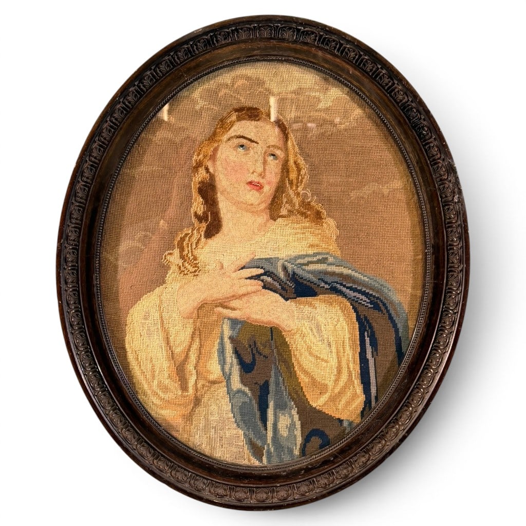 Oval Religious Needlepoint: Oval framed needlepoint panel depicting a religious female figure, likely the Virgin Mary or a saint, shown with blue mantle and clasped hands, mounted under glass in an oval carved dark wood frame.