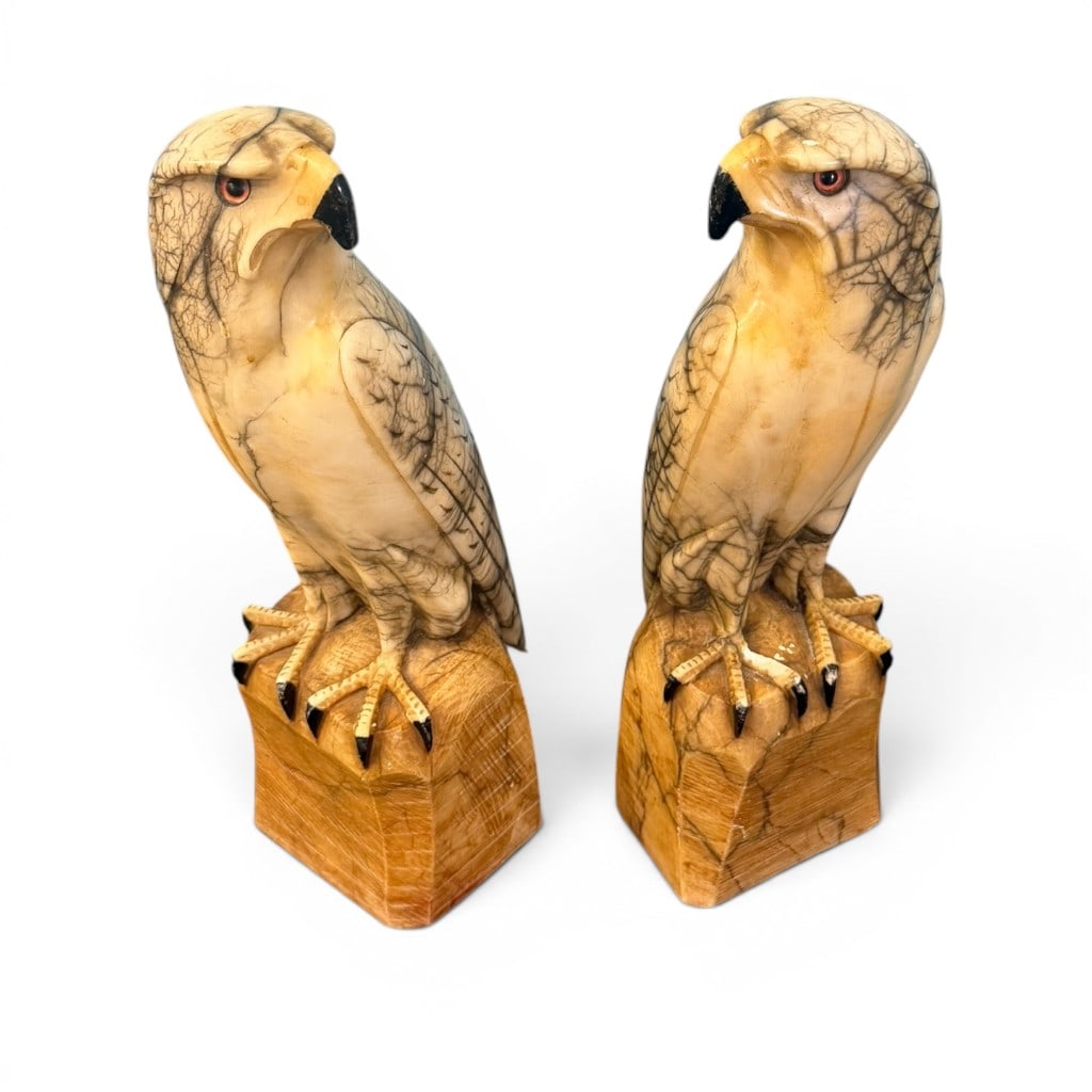 Pair of Carved Marble Falcon Figures on Bases: Pair of carved marble falcon figures, each modeled perched on an integral block base with black-painted beak and talons and inset red eyes. The birds are carved from variegated cream, tan, and gray-ve