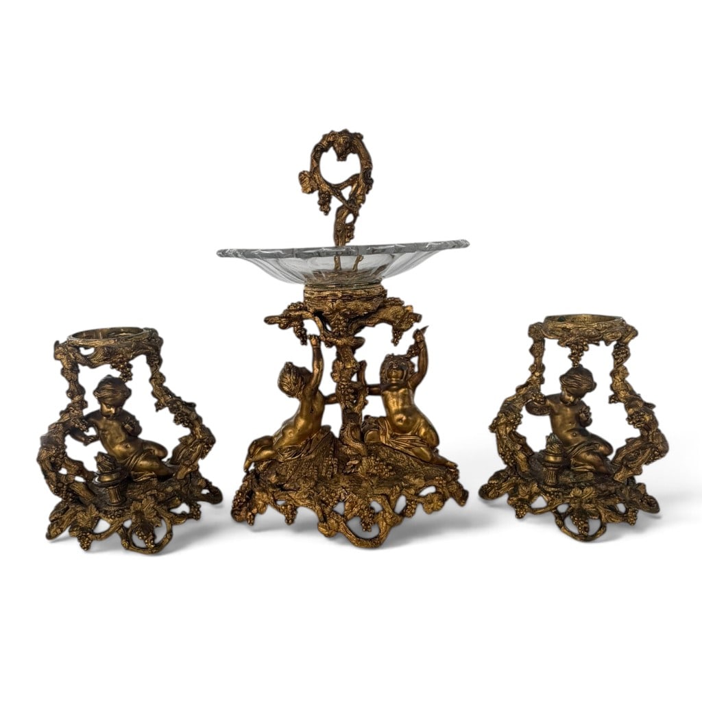 Fine Three-Piece Gilt-Metal Putti Garniture: Origin: Continental Europe, probably France Date: Late 19th to early 20th century Period/Style: Rococo Revival Maker: Unidentified Materials: Gilt metal and pressed glass Form: Three-piece tabl