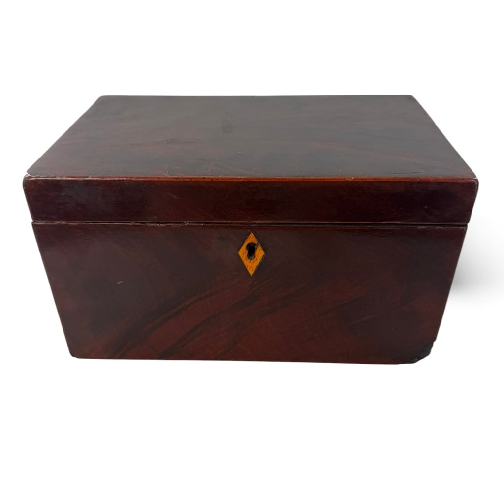 Regency Mahogany Two-Compartment Tea Caddy: Origin: England. Date: Circa 1820-1840. Description: English figured mahogany tea caddy of rectangular form with subtly canted sides and a diamond-form lightwood escutcheon. The hinged lid has a red t