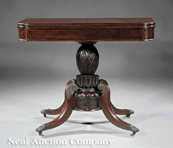 An American Classical Carved Games Table: An American Classical Carved Games Table, early 19th c., Boston, foldover top, pineapple-carved standard, drum plinth, four outswept legs with boss terminals, paw cuffs, casters, height 29 in., width 