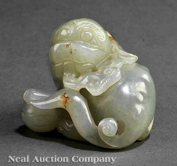 Chinese Celadon Jade Buddhist Lion: A Chinese Celadon Jade Buddhist Lion, 19th c., carved reclining on its side with a ball beneath its forepaws and a lingzhi spray in its mouth, having a distinct ridged backbone, height 1 3/8 in., widt