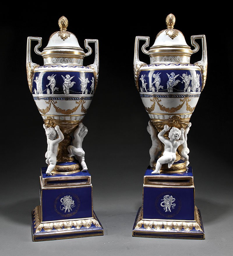 A Pair of Elaborate Minton-Style Porcelain Urns: A Good Pair of Elaborate Minton-Style Porcelain Urns, with covers, the large urn-form vases fictively supported by three bisque putti, on tall cobalt glazed plinths, decorated in thin grisaille relief