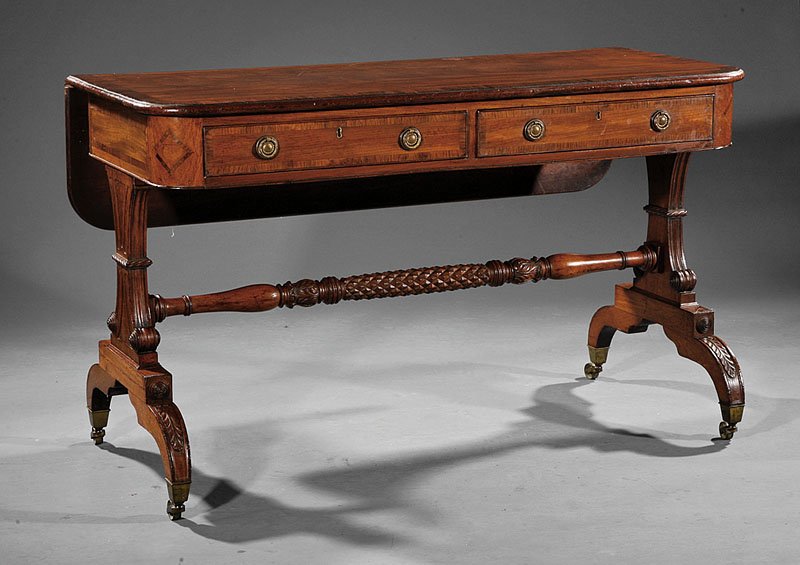 A George IV Carved Mahogany and Rosewood Banded S: A George IV Carved Mahogany and Rosewood Banded Sofa Table, c. 1825, D shaped top with a drop leaf, conforming frieze with two drawers, trestle supports with bosses, turned stretcher, acanthine arched