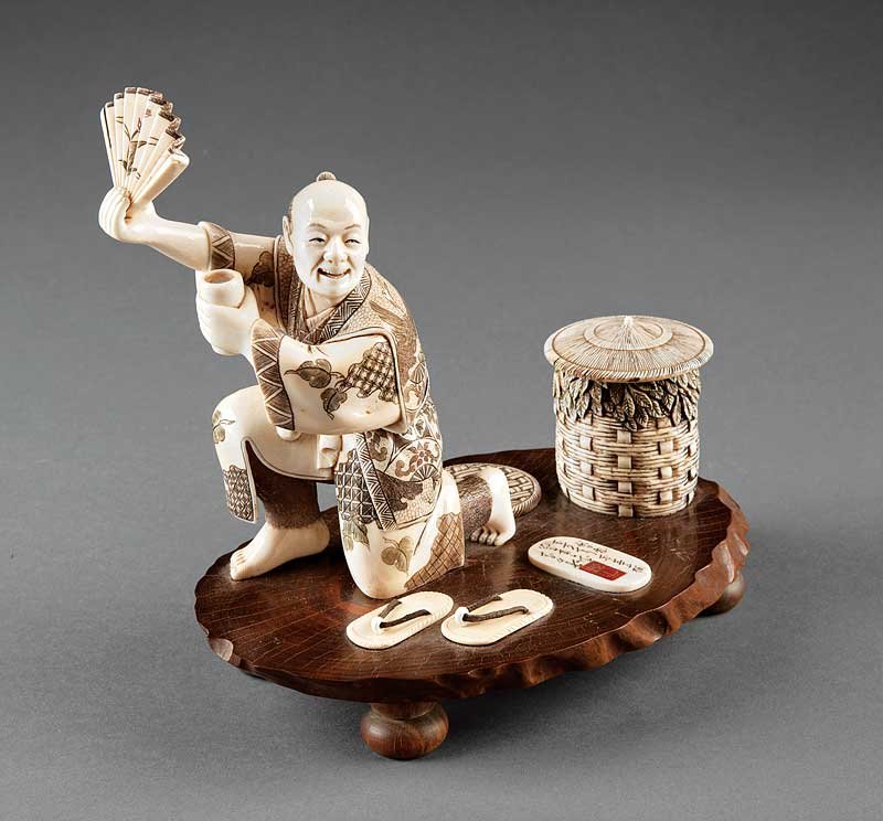 An Japanese Ivory Figural Group: An Antique Japanese Ivory Figural Group, carved as a man holding a fan and a cup kneeling beside a covered basket and a pair of sandals, height 6 3/8 in., wood base inset with inscribed ivory cartouch