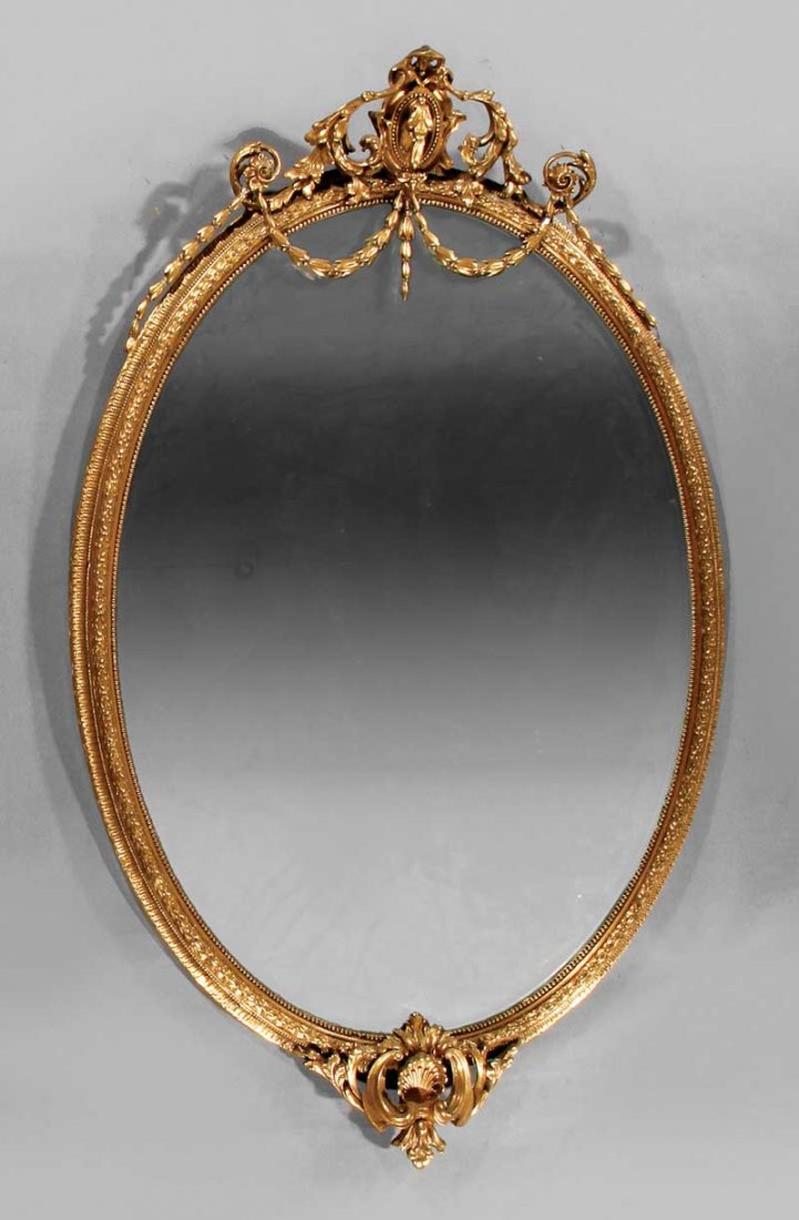 Carved Giltwood Mirror stamped John Taylor & Sons: A Fine Scottish Carved Giltwood Mirror, in the Louis XVI taste, stamped John Taylor & Sons, Manufacturer, Edinburgh, oval bellflower swagged frame surmounted by a beaded cartouche, base with foliate s