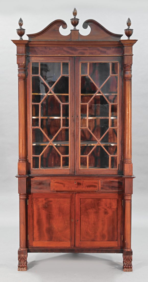 An American Carved Mahogany Corner Cabinet: An American Carved Mahogany Corner Cabinet, 19th c., upper case with broken arch crest and urn finials, blocked frieze, glazed doors, shelf interior, flanking columns; conforming lower case with a med