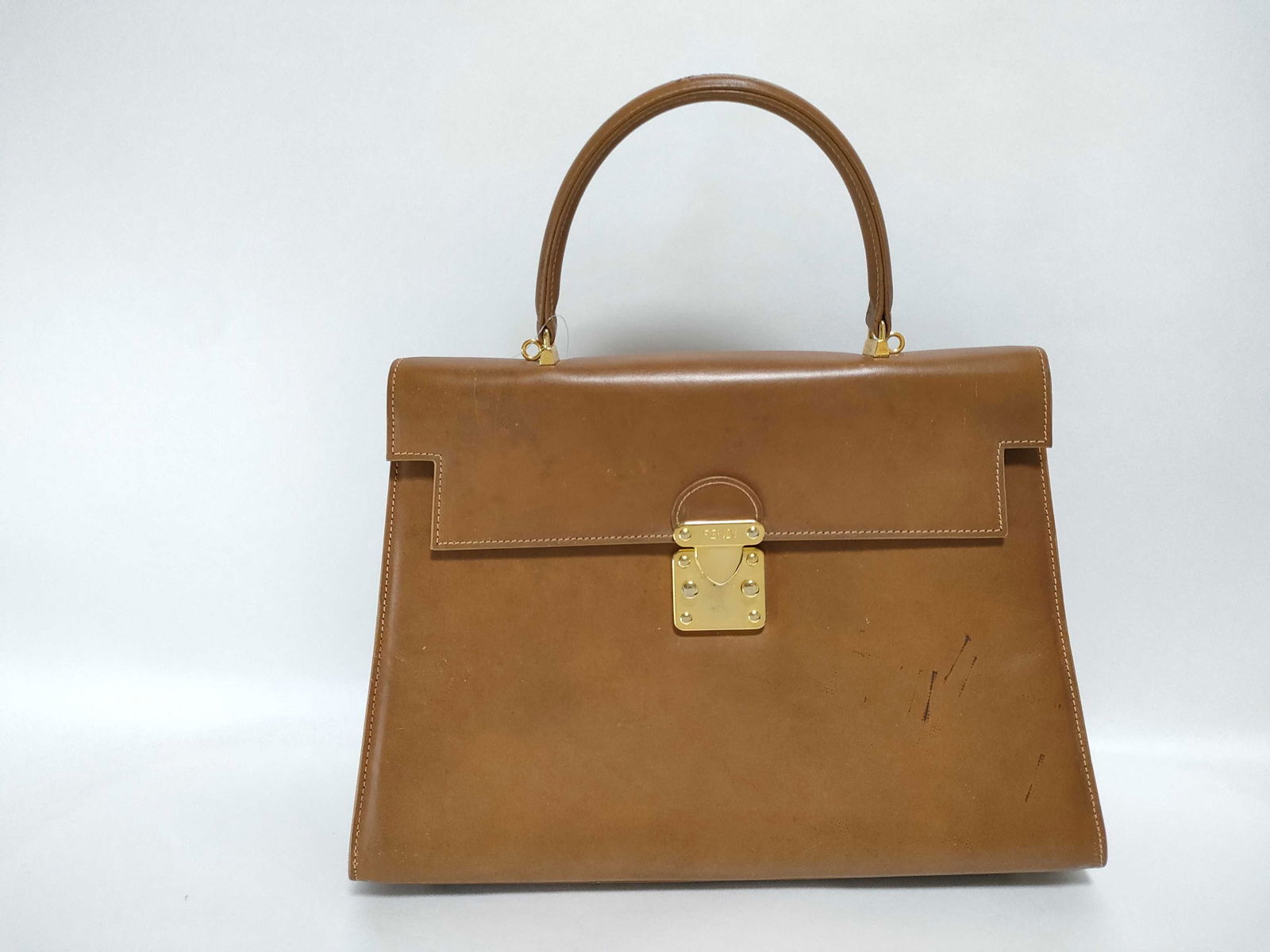 Fendi Tan Leather Top Handle Handbag with Lock Hardware: fendi handbag, authentic. made of smooth tan leather with structured trapezoid shape. single top handle attached by gold-tone hardware. front flap with rectangular gold lock closure centered near top