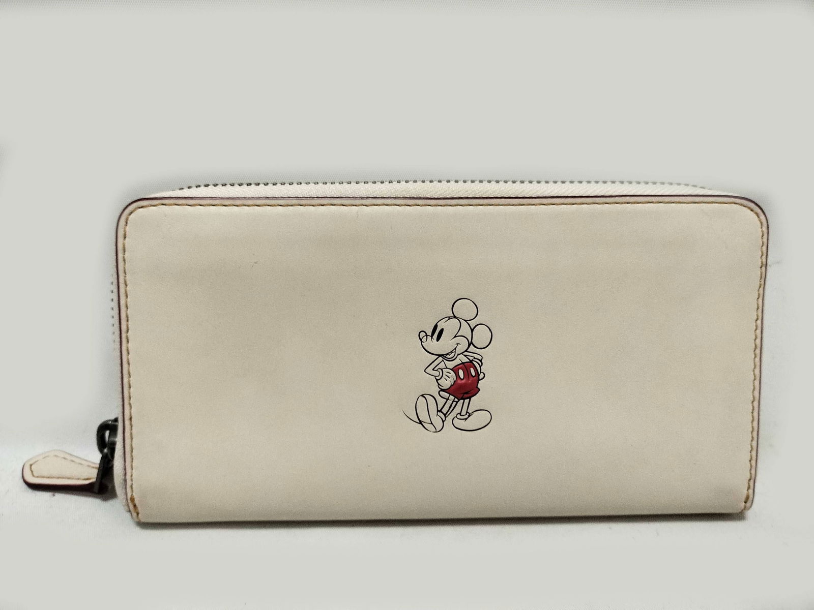 Coach x Disney collaboration long zip-around wallet: Coach x Disney collaboration long zip-around wallet in white leather featuring printed Mickey Mouse graphic in red shorts and yellow shoes on front. Silver-tone zipper closure with leather pull tab. I