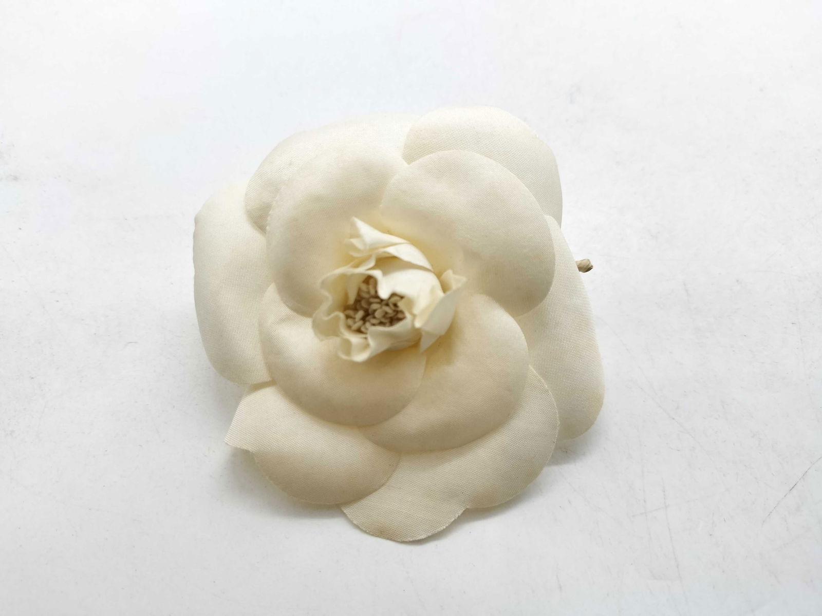 Chanel Camellia Corsage Brooch with box: White fabric camellia with layered petals forming rounded bloom. Central stamen detail in matching tone. Metal pin backing with straight bar and safety clasp. Gold-tone oval Chanel plate marked “Mad