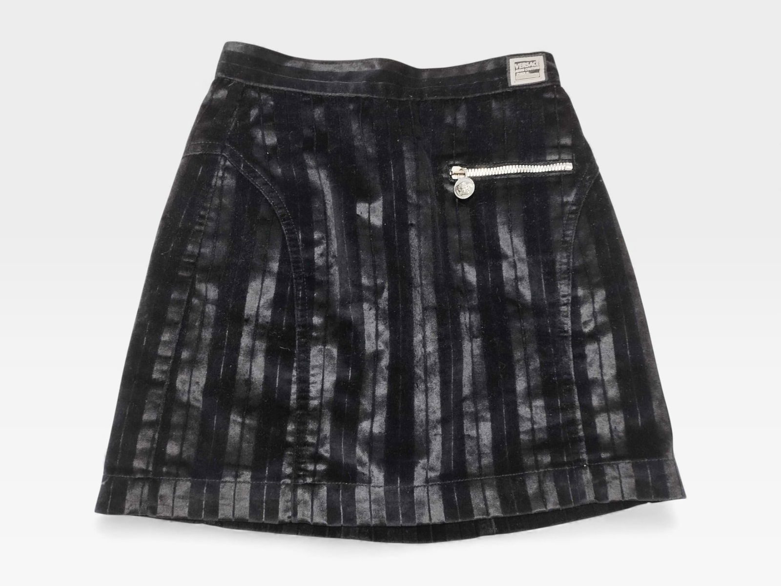 Versace Black Velour Mini Skirt with Medusa Hardware: Black velour mini skirt with vertical stripe texture and subtle sheen. Front zip pocket with silver-tone Medusa pull. Side and back seams shaping slight A-line silhouette. Back zipper and button closu