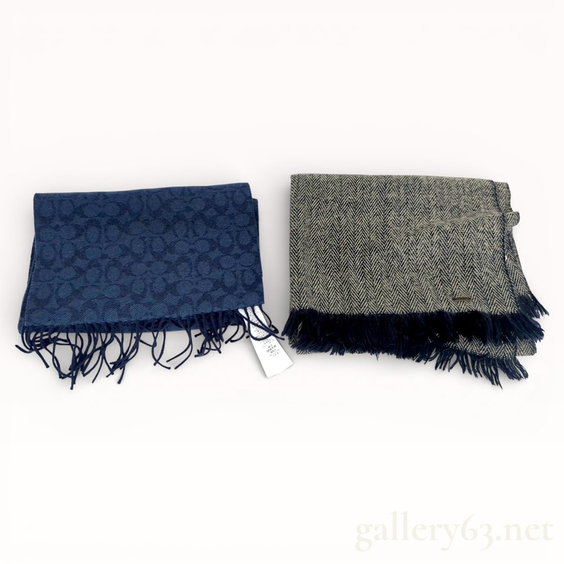 Designer Muffler Pair Including Coach: Group of two muffler scarves comprising one example by Coach and one by another maker. Set includes a blue patterned muffler with fringed ends and a gray herringbone-patterned muffler finished with