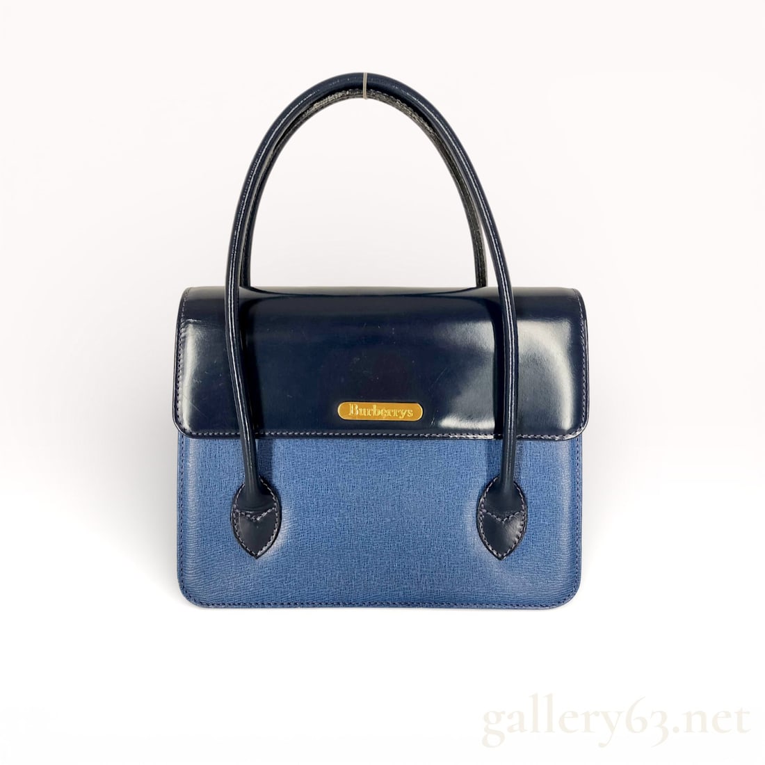 Burberry Blue Label Blue and Navy Handbag: Authentic Burberry Blue Label handbag featuring a structured silhouette in blue canvas with navy leather trim. Dual top handles and flap-style top with gold-tone logo plaque. Compact, structured form