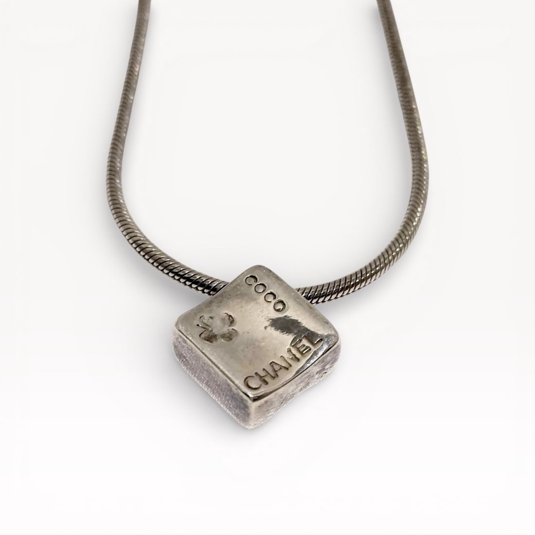 Chanel Camellia Necklace in Sterling Silver with Logo Pendant: Authentic Chanel Camellia necklace in sterling silver featuring a cube-shaped pendant engraved with the Chanel name and camellia flower motif. Pendant suspended from a flexible sterling silver snake