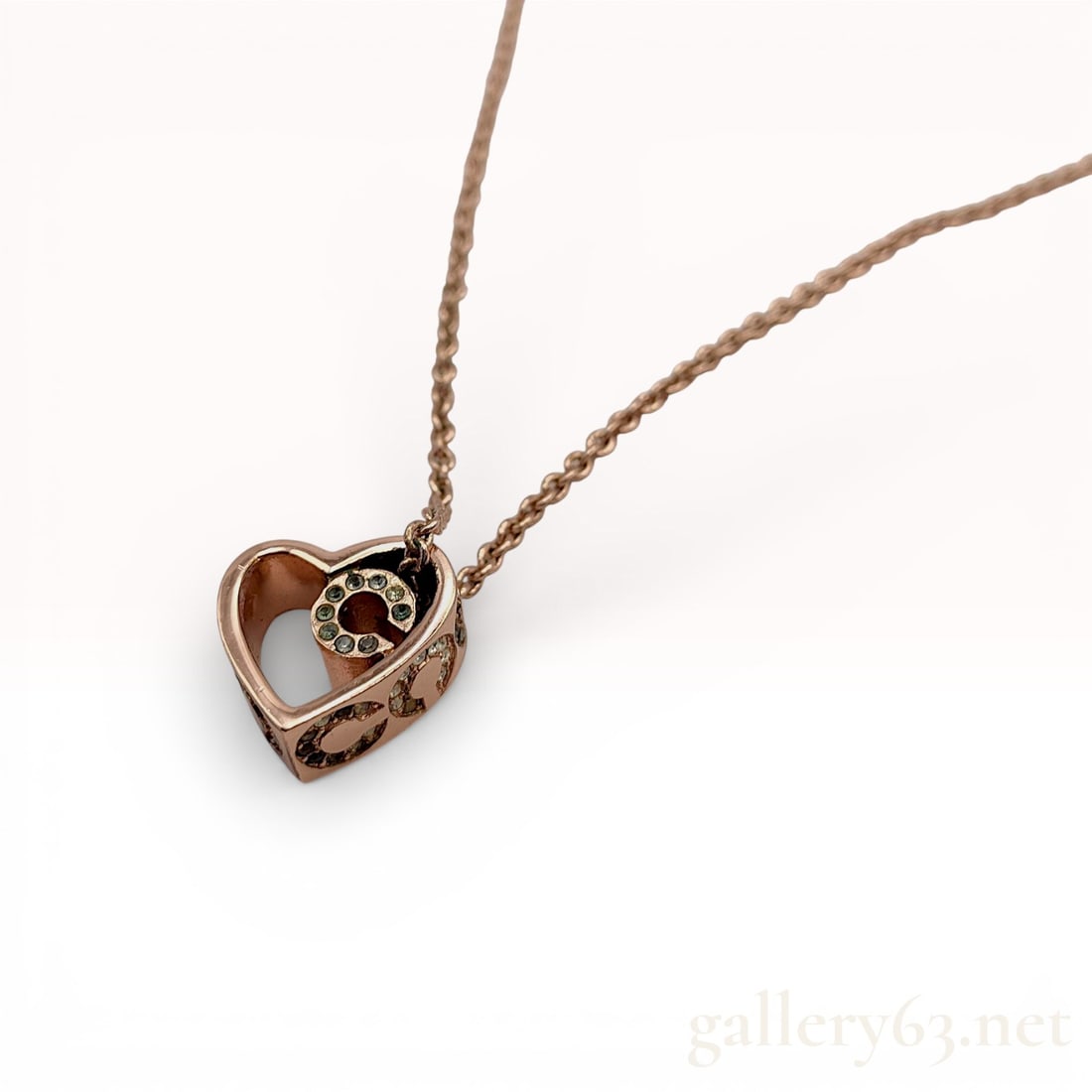 Coach Rose Gold-Tone Rhinestone Heart Pendant Necklace: Coach necklace featuring a rose gold-tone heart-shaped pendant with cutout detailing. Design accented by rhinestone-set logo elements within the heart motif. Pendant suspended from a fine chain wi