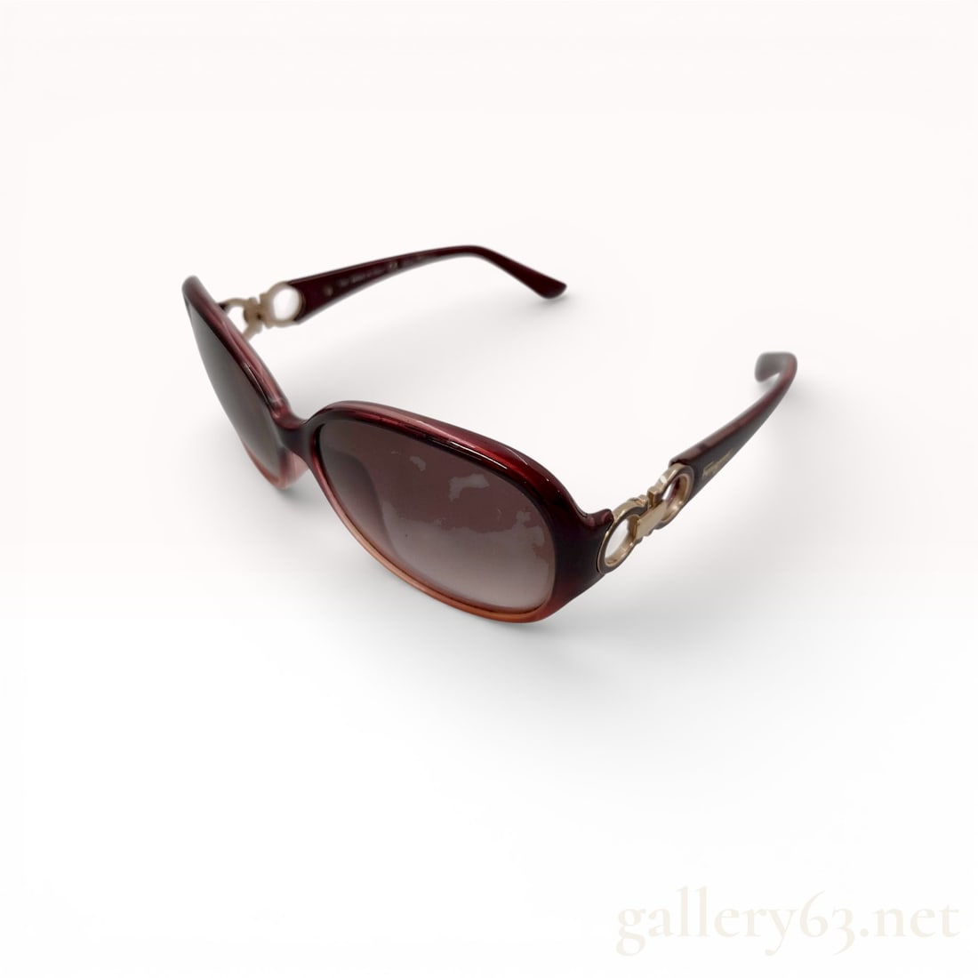Salvatore Ferragamo Brown Gradient Sunglasses with Gold-Tone Gancini Temple Accents: Salvatore Ferragamo sunglasses featuring brown translucent frames in a softly rounded silhouette. Design accented by gold-tone Gancini-style hardware at the temples. Lenses finished in a dark grad