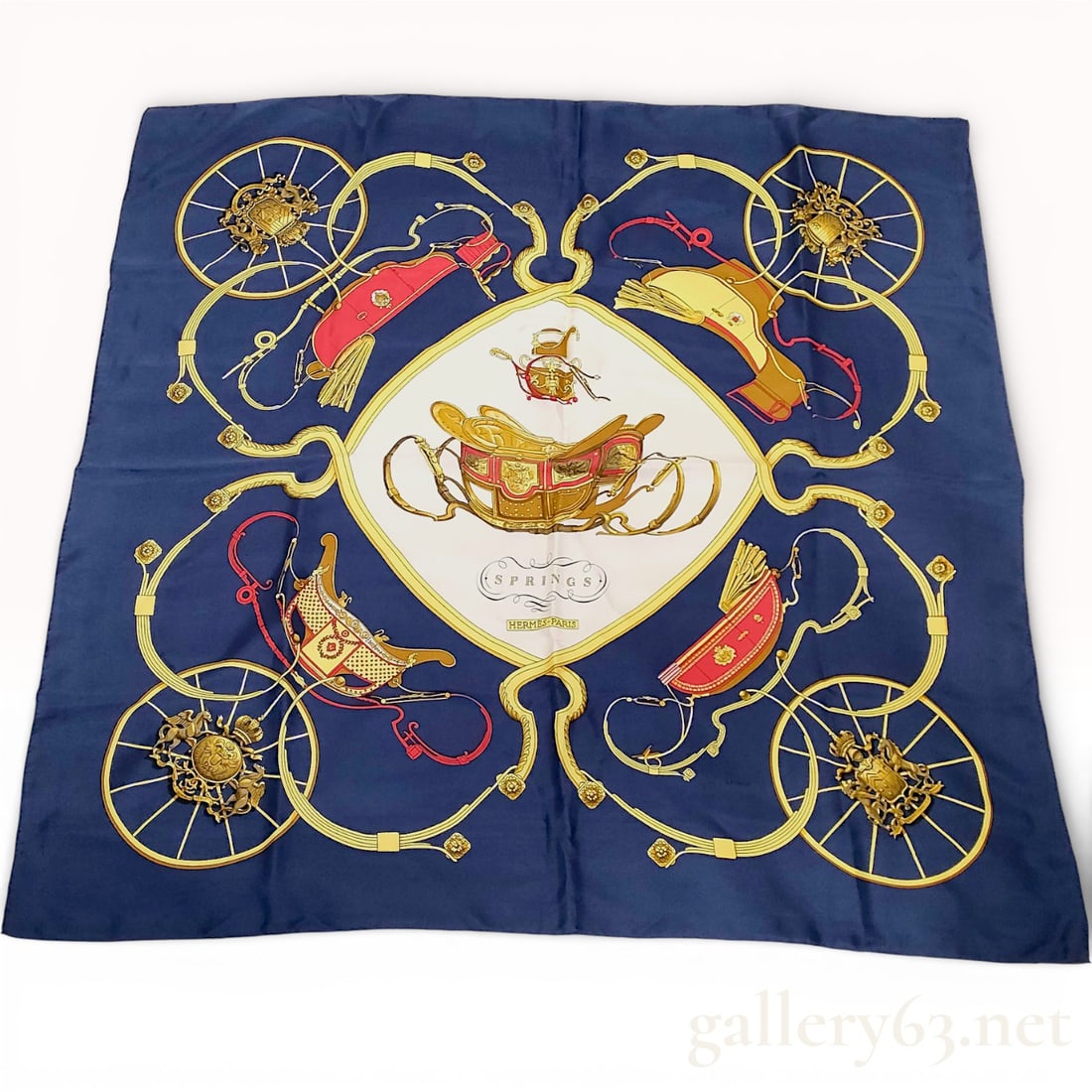 Hermès “Springs?? Carré 90 Silk Scarf in Navy with Carriage and Wheel Motif: Hermès “Springs” carré 90 silk scarf featuring a central carriage motif framed by wheels, harness straps, and tassel elements in gold, red, and cream tones against a navy ground.