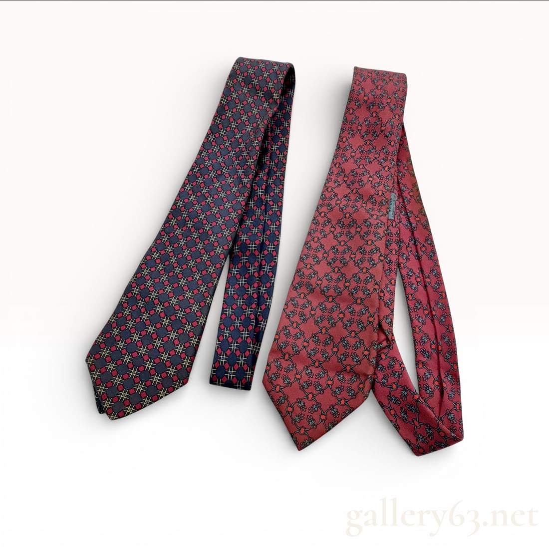 Two Hermes Silk Neckties: Two Hermes silk neckties. The first is a navy blue silk tie featuring a repeating geometric pattern in red and gold. It includes a woven label with the Hermes horse and carriage logo and dry clean onl