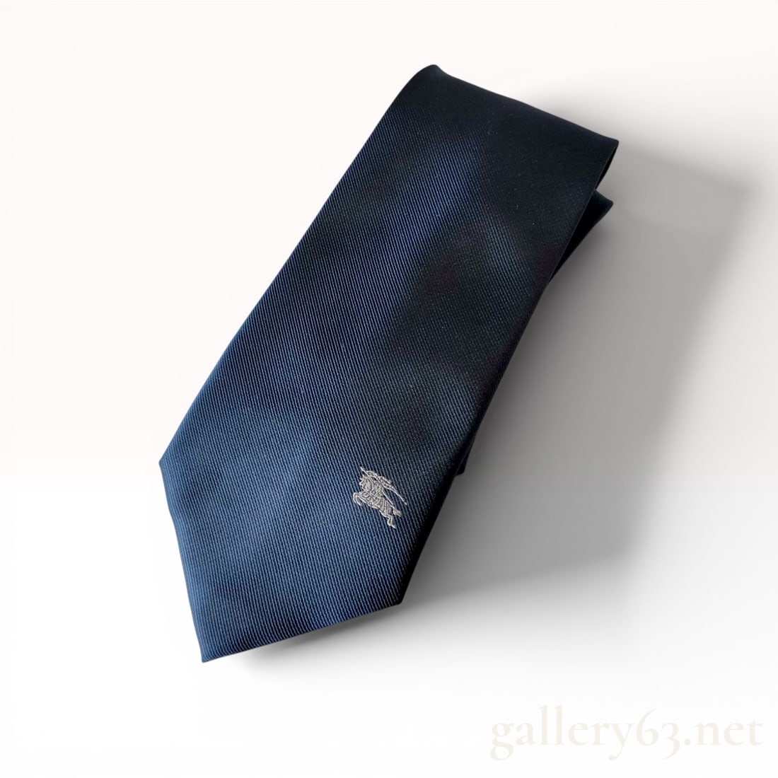 Burberry London Navy Blue Silk Prorsum Knight Logo Necktie: Burberry London necktie in solid navy blue silk featuring a diagonal twill weave. The blade is detailed with a silver-tone embroidered Prorsum Knight logo, while the narrow end features a square-frame