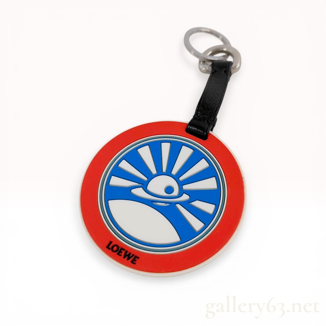 Loewe Eye and Sunburst Graphic Rubber and Leather Keychain: Loewe keychain featuring a circular rubber fob with a red, blue, and white stylized eye and sunburst graphic. The fob is attached to a black leather strap with silver-tone metal hardware including a s