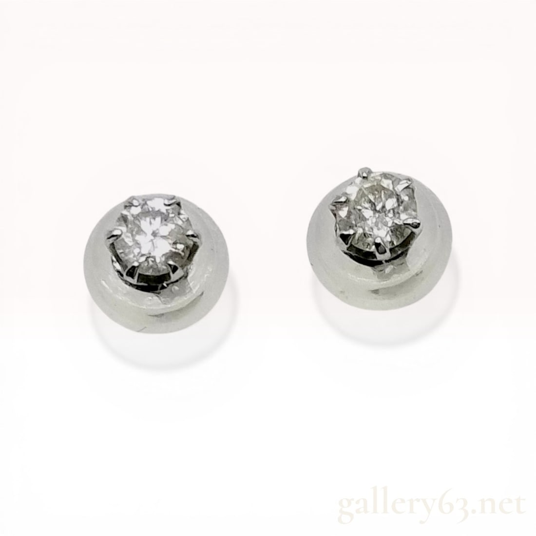 A Pair of Diamond and White Metal Six-Prong Stud Earrings: A matched pair of stud earrings featuring two round brilliant-cut diamonds. Each stone is secured in a six-prong basket setting of polished white metal with post and friction backings. The posts are i