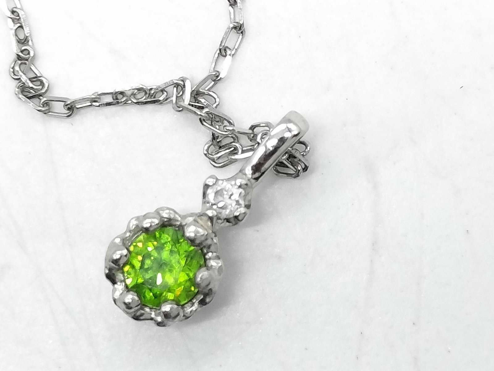 Platinum Peridot and Diamond Pendant Necklace: Platinum pendant necklace featuring a round faceted green peridot and a small clear accent diamond. The pendant is constructed of Pt900 platinum with a textured prong setting, suspended from a fine Pt