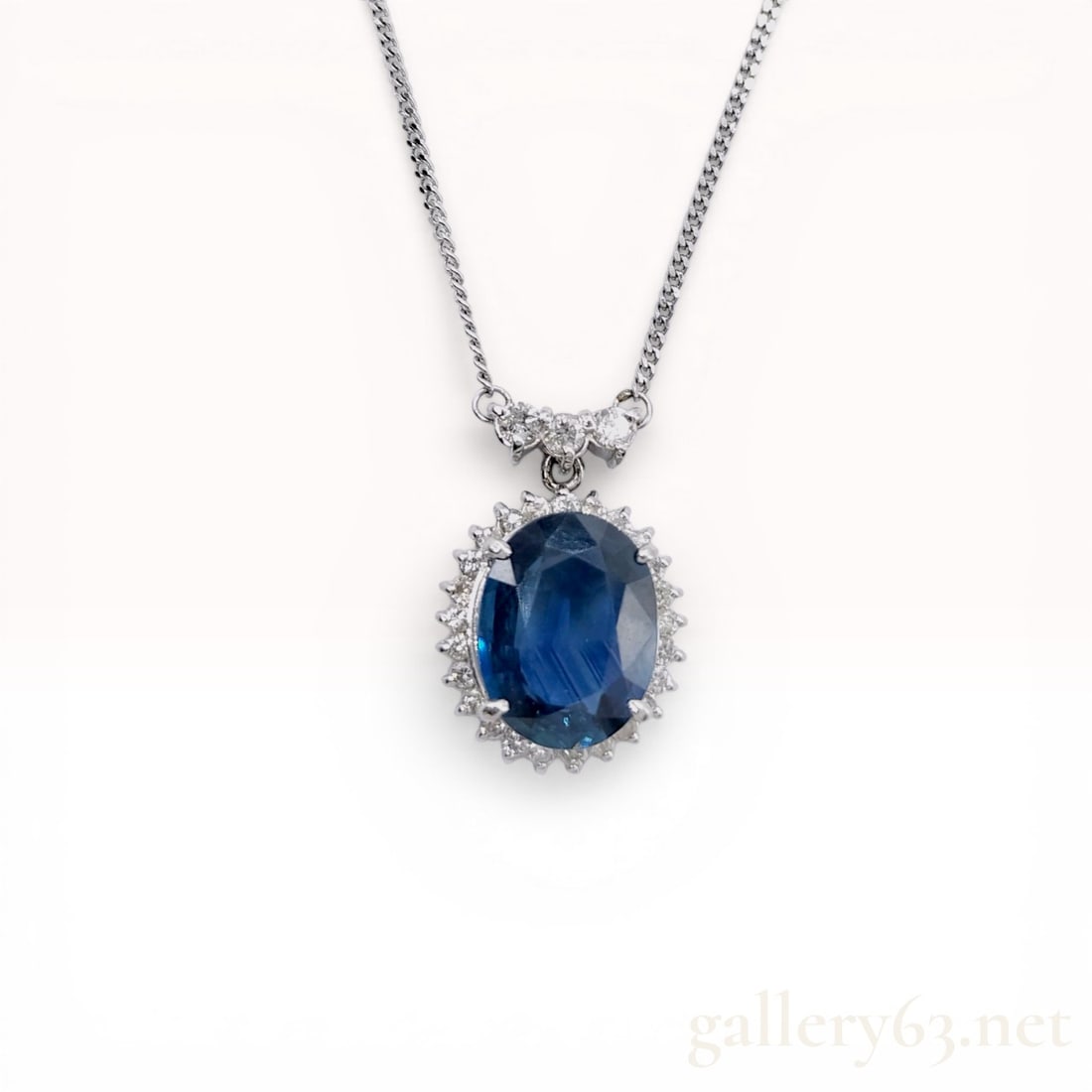 Platinum Sapphire and Diamond Halo Pendant Necklace: Platinum pendant necklace featuring a central oval-cut blue sapphire. The sapphire is bordered by a halo of round brilliant-cut diamonds and hangs from a stationary bail accented with three graduated