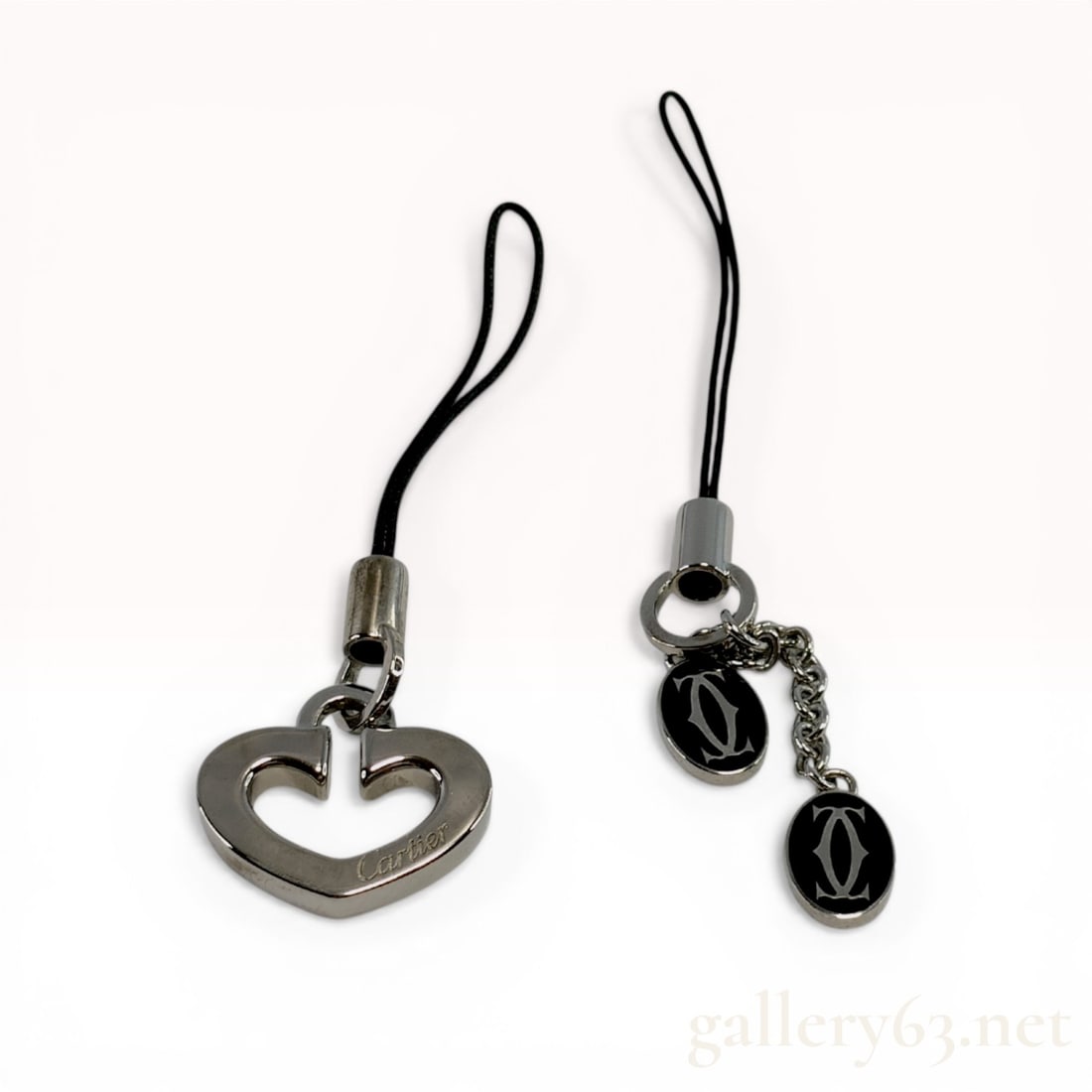 Two Cartier Silver-Tone Mobile Phone Straps: Cartier accessory charms, early 21st century. One charm features a stylized open heart with an incised cursive Cartier signature, while the other features two oval medallions with black enamel-style f