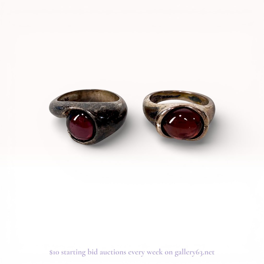 A Pair of Modernist Silver and Garnet-Colored Cabochon Rings: A pair of modernist silver fashion rings featuring round garnet-colored cabochon stones. Each ring is constructed with a bypass or swirl setting where the metal band wraps around the central stone. Th