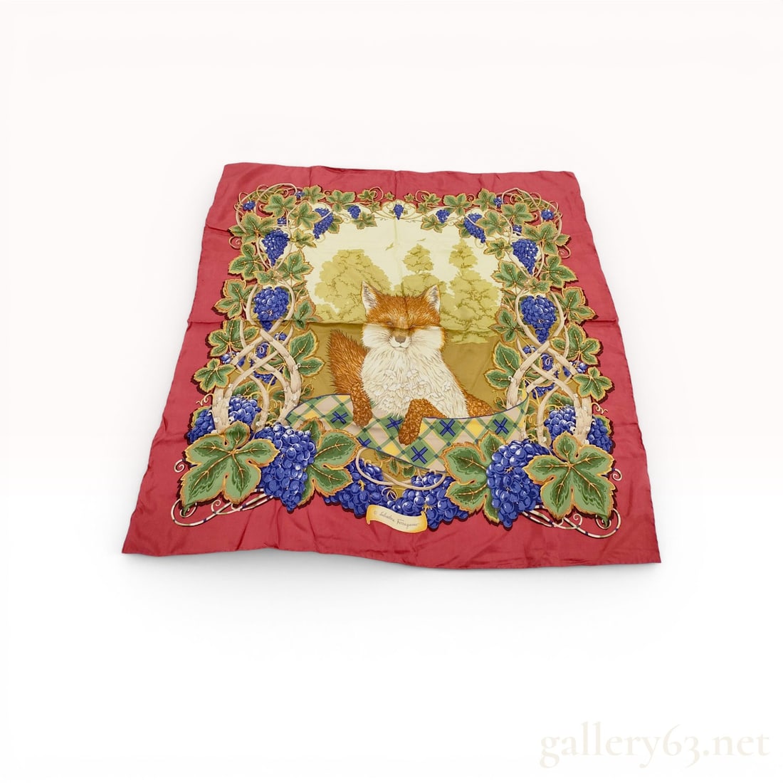 Salvatore Ferragamo Silk Scarf in Fox and Grapes Pattern: A square silk foulard by Salvatore Ferragamo featuring a central illustration of a red fox resting on a yellow, green, and blue plaid blanket. The central image is set against a pale yellow landscape 