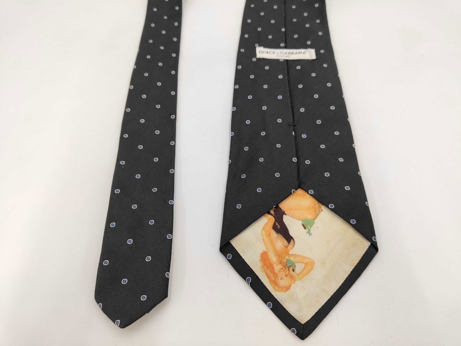 Dolce & Gabbana Silk Pin-Dot Tie with Pin-Up Graphic Lining: Dolce & Gabbana 100% silk necktie, made in Italy. Features an embroidered blue and grey pin-dot pattern on a black ground with a hidden vintage-style pin-up girl graphic on the interior tipping. I