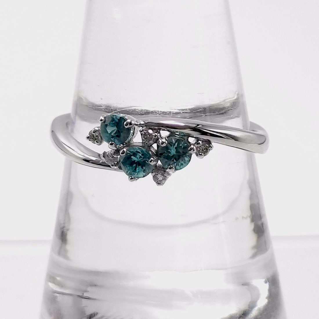 18K White Gold Teal-Blue Sapphire and Diamond Bypass Cluster Ring: This 18 karat white gold ring features a contemporary bypass or wave design. The central cluster is composed of three round-cut teal-blue sapphires, complemented by seven small round-cut white diamond