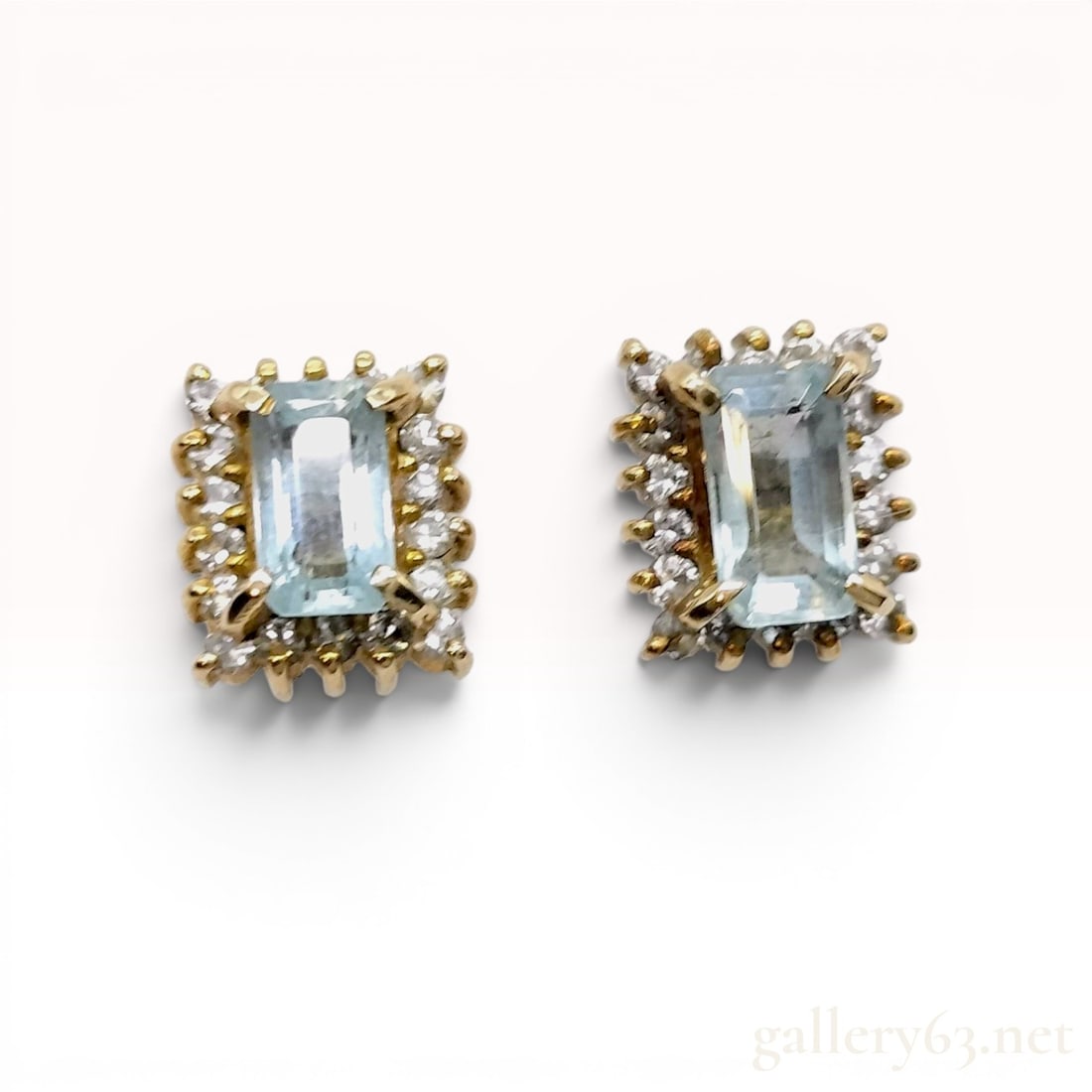 A Pair of Aquamarine and Diamond Halo Stud Earrings in 18K Yellow Gold: Matched pair of 18-karat yellow gold stud earrings featuring a classic halo cluster design for pierced ears. Each earring is set with a central emerald-cut aquamarine framed by a border of small round