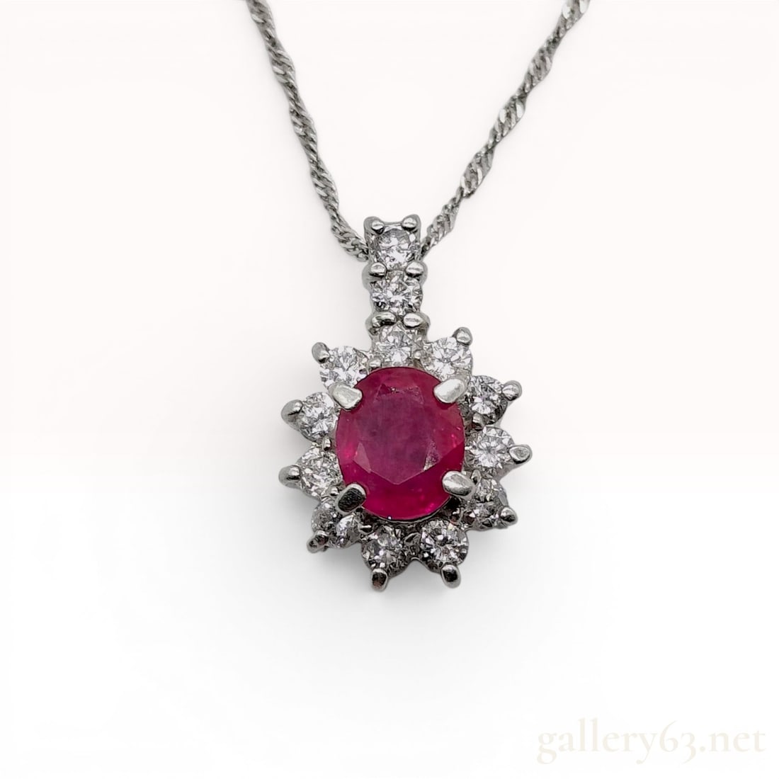 Platinum 900/850 Ruby and Diamond Cluster Pendant Necklace: A platinum and ruby cluster pendant suspended from a platinum chain. The pendant features a central prong-set oval-cut red ruby, measuring 0.40 carats. Surrounded by a halo of diamonds. The necklace u