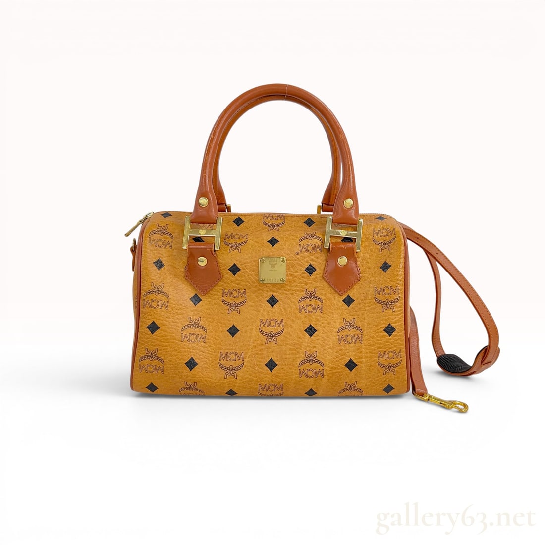 MCM Cognac Monogram Visetos Boston Bag with Shoulder Strap: An MCM Boston-style satchel crafted from the signature Cognac Visetos monogram coated canvas with tan leather trim. The exterior features the repeating MCM laurel logo and text print. The bag is desig