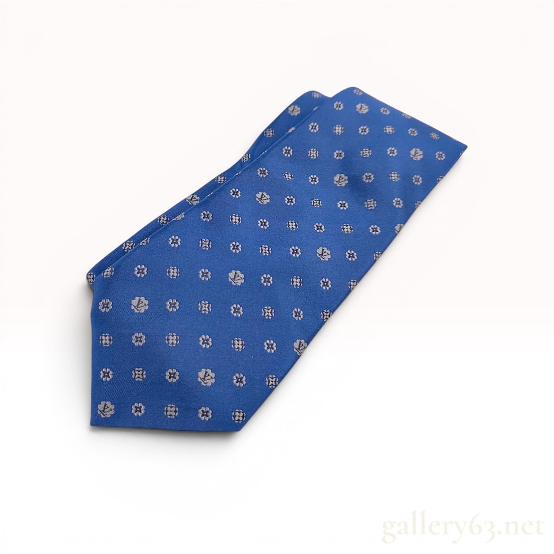 Louis Vuitton Monogram Floral Silk Necktie: Louis Vuitton men's necktie featuring a repeating silver-toned floral and monogram jacquard pattern on blue silk. The narrow end displays a distinct square embroidered LV monogram, while the reverse i