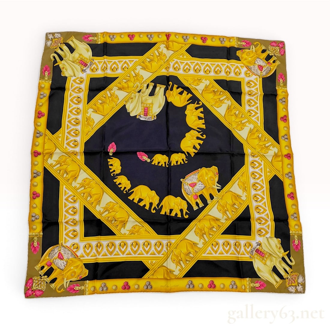 Must de Cartier L'Elephant Pattern Square Silk Scarf: A square silk scarf from the Must de Cartier line featuring the L'Elephant motif. The design centers on a navy blue field with a spiraling procession of golden elephants. This central section is frame