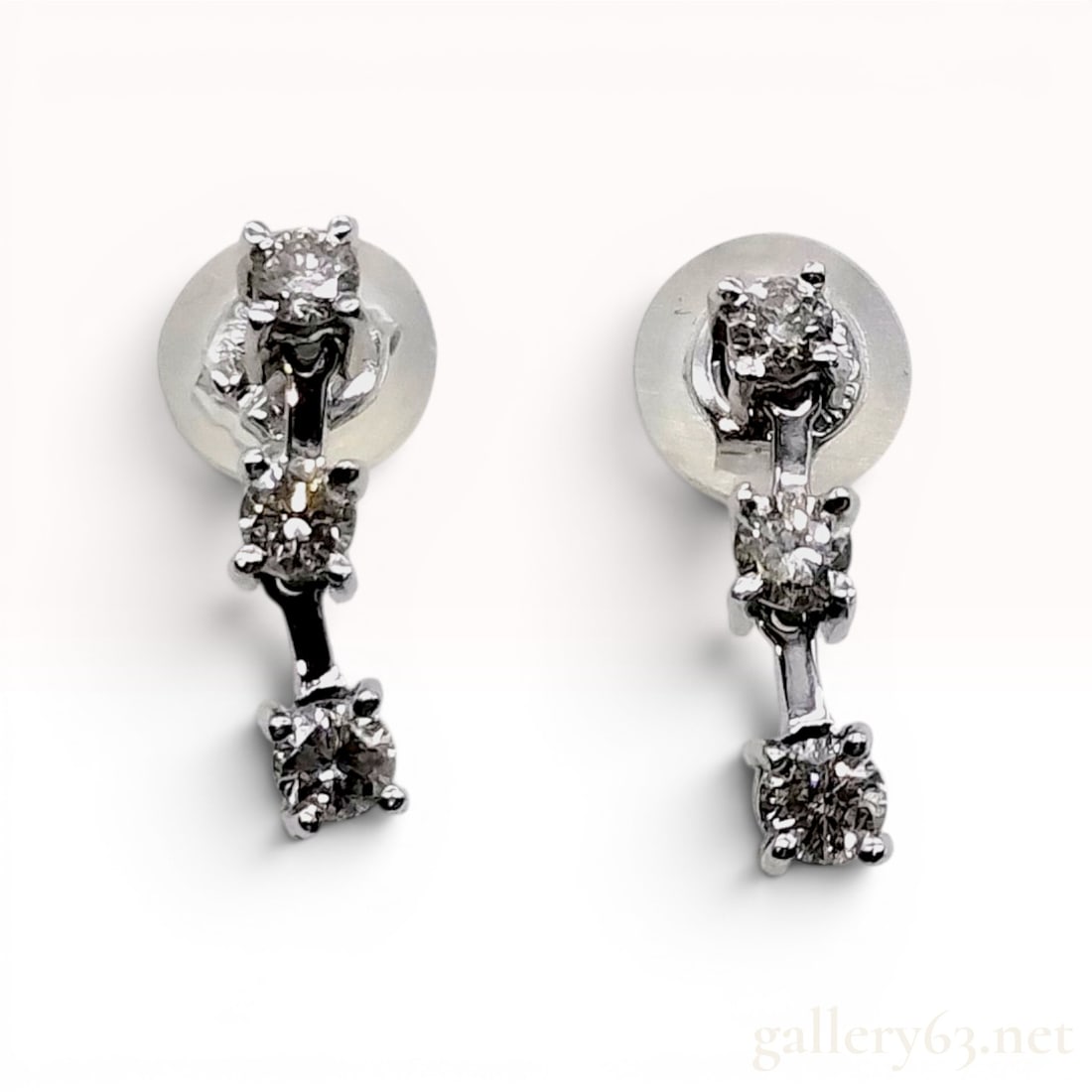 A Pair of 18K White Gold Three-Stone Diamond Linear Drop Earrings: A matched pair of linear three-stone drop earrings crafted in 18-karat white gold. Each earring features three round brilliant-cut diamonds in graduated sizes, individually secured within four-prong s