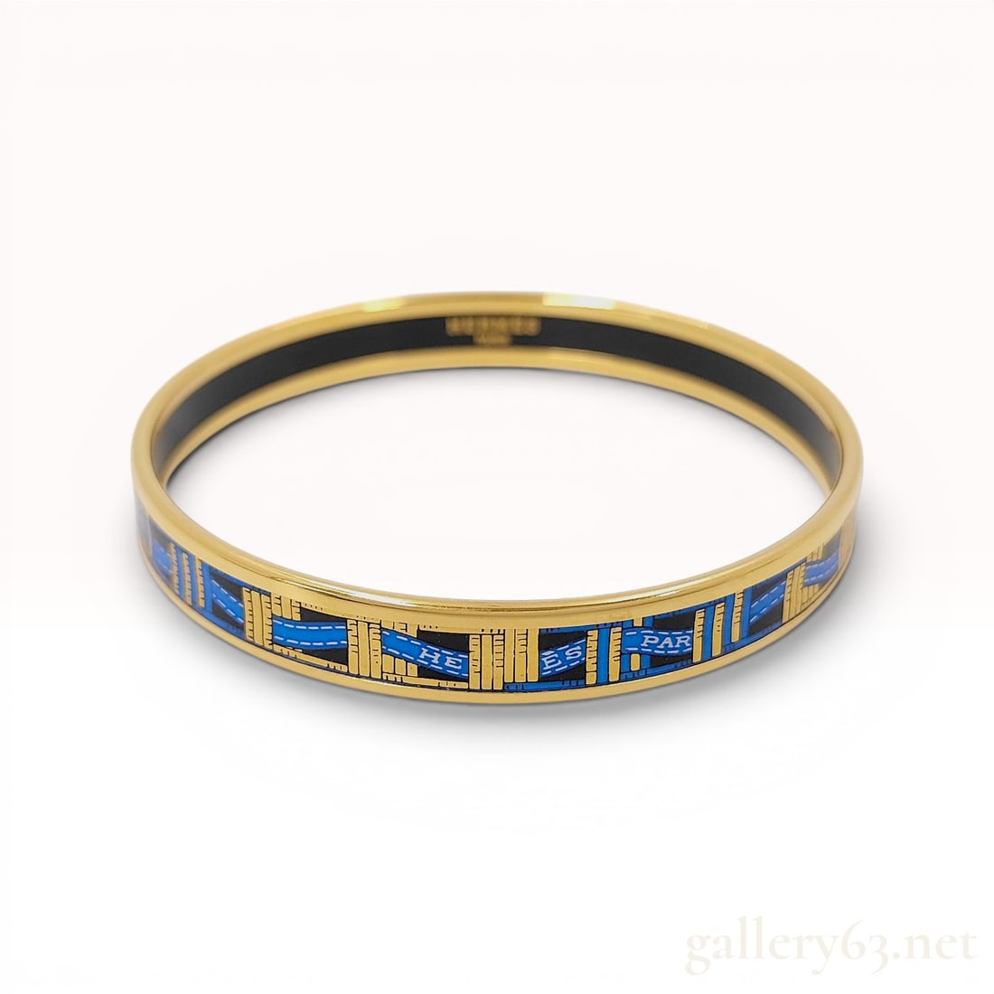 Hermes Gold-Tone and Enamel Narrow Ribbon Motif Bangle Bracelet: A Hermes narrow enamel bangle bracelet. The jewelry item is constructed from gold-tone metal featuring a printed enamel inlay. The exterior design displays a pattern of blue ribbons inscribed with the