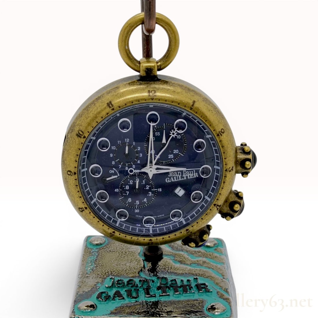Jean Paul Gaultier Alarm Chronograph Pocket Watch with Presentation Stand Reference 0580-H33171: This Jean Paul Gaultier pocket watch and stand set features an antiqued brass-tone finish over a stainless steel case. The navy blue dial is configured with a multi-functional chronograph layout compr