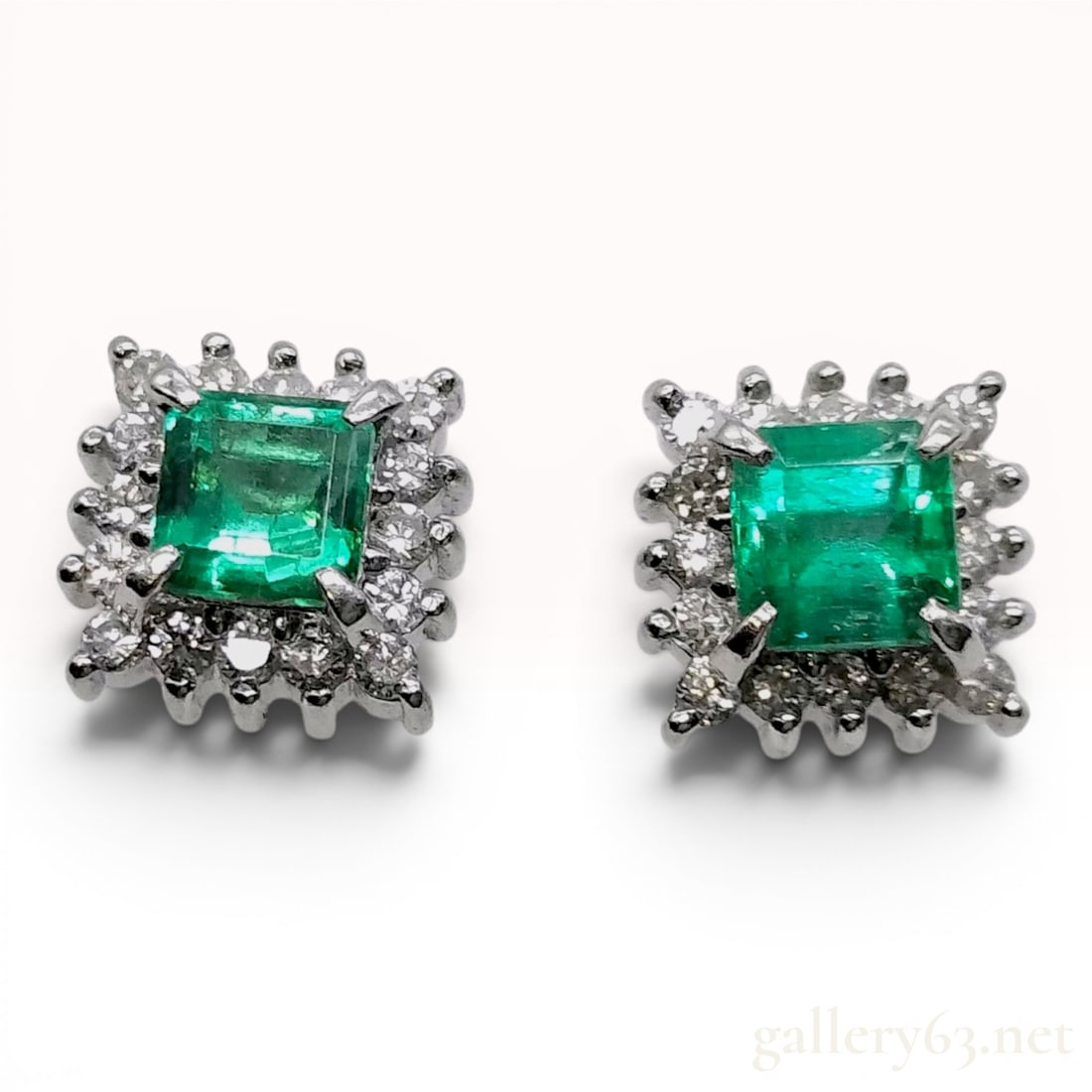 A Pair of Emerald and Diamond Platinum Cluster Stud Earrings: A matched pair of platinum cluster stud earrings in a square halo design. Each earring is centered with a square step-cut green emerald in a four-prong setting. The emerald is bordered by a halo of tw
